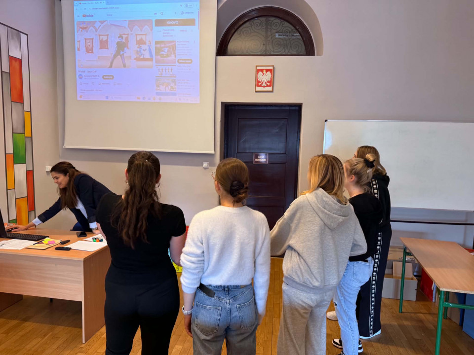Na zdjęciu w sali zajęciowej pięcioro studentów stoi w kółku czekając na instrukcję od nauczyciela. Nauczyciel przy biurku próbuje coś zrobić na laptopie.